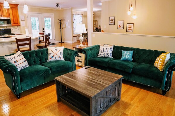 Una sala de estar con dos sofás de terciopelo verde, una mesa de centro de madera y una cocina al fondo.