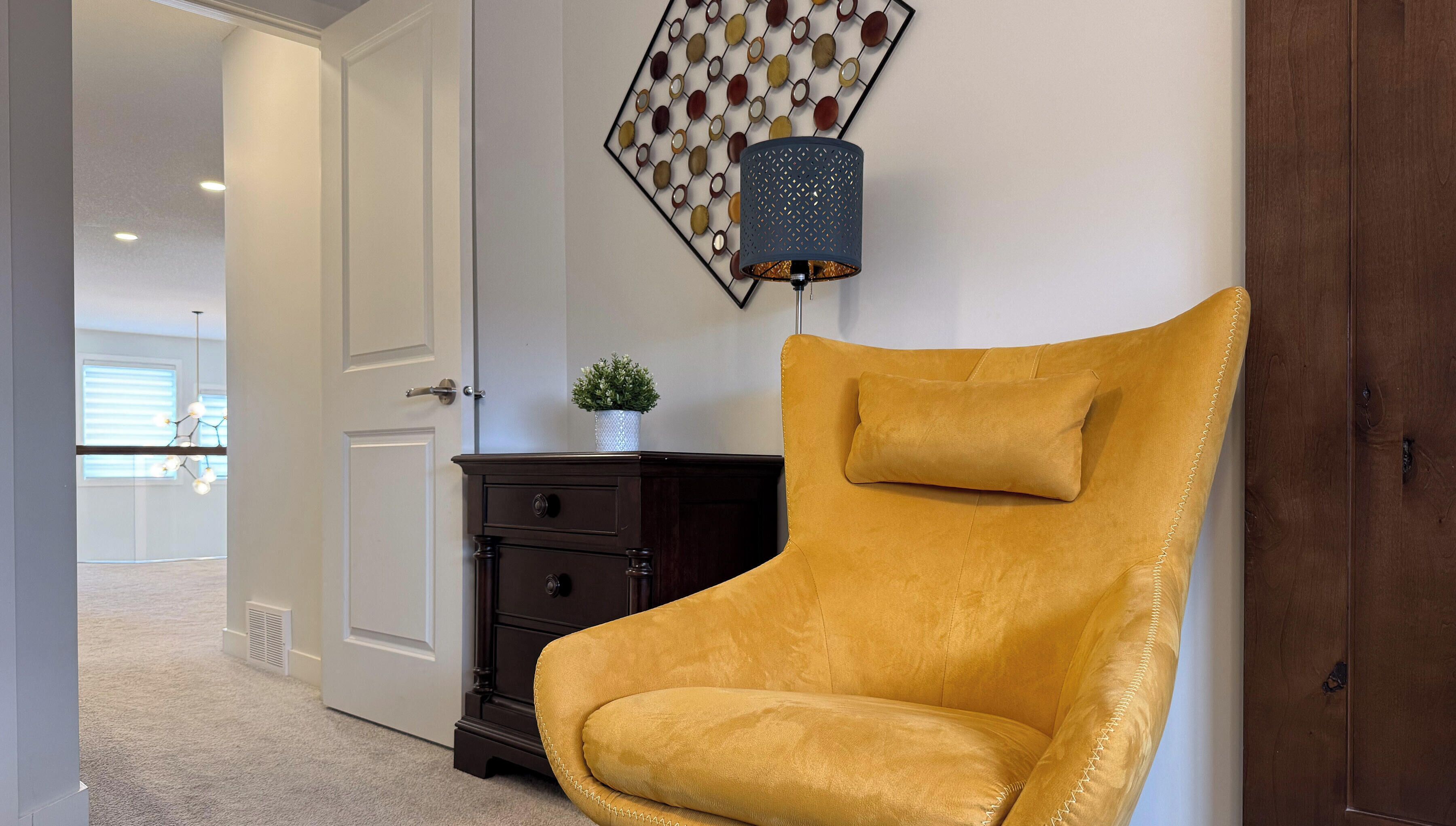 Een mosterdkleine fauteuil met een hoge rugleuning en een kussen, geplaatst voor een donkere houten kast en een decoratief wandobject.