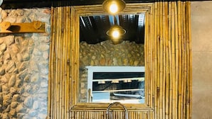 A bathroom with a stone wall, a wooden shelf with toiletries, and a mirror framed by bamboo.