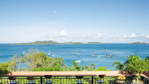 A seaside view with a wooden deck and chairs overlooking the ocean, boats, and distant hills.