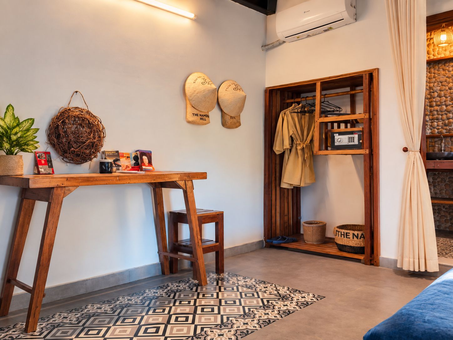 Una habitación acogedora con un escritorio de madera, un tapete estampado, un taburete pequeño, un estante con ropa y un aire acondicionado fijado a la pared.