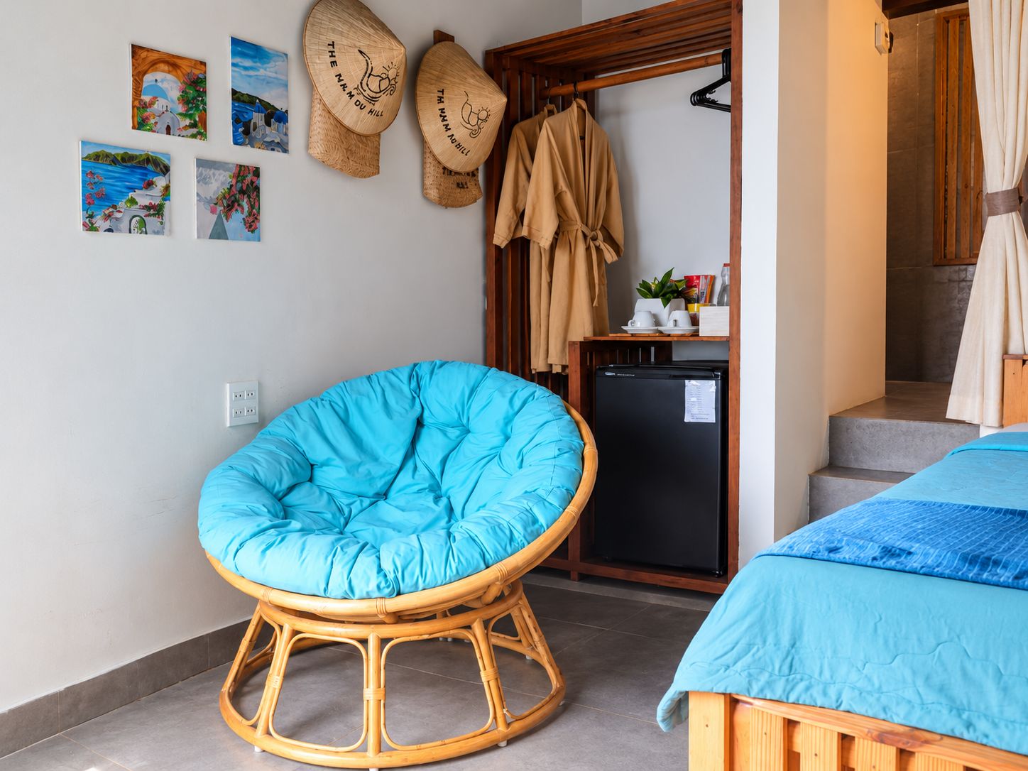 Une pièce avec un fauteuil bleu rembourré, un lit recouvert d’une courtepointe bleue, un petit réfrigérateur noir et une porte en bois sur laquelle est accrochée une serviette.