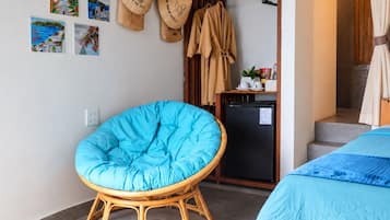Una habitación con una silla tapizada en azul, una cama con colcha azul, un refrigerador negro pequeño y una puerta de madera con una toalla colgada.