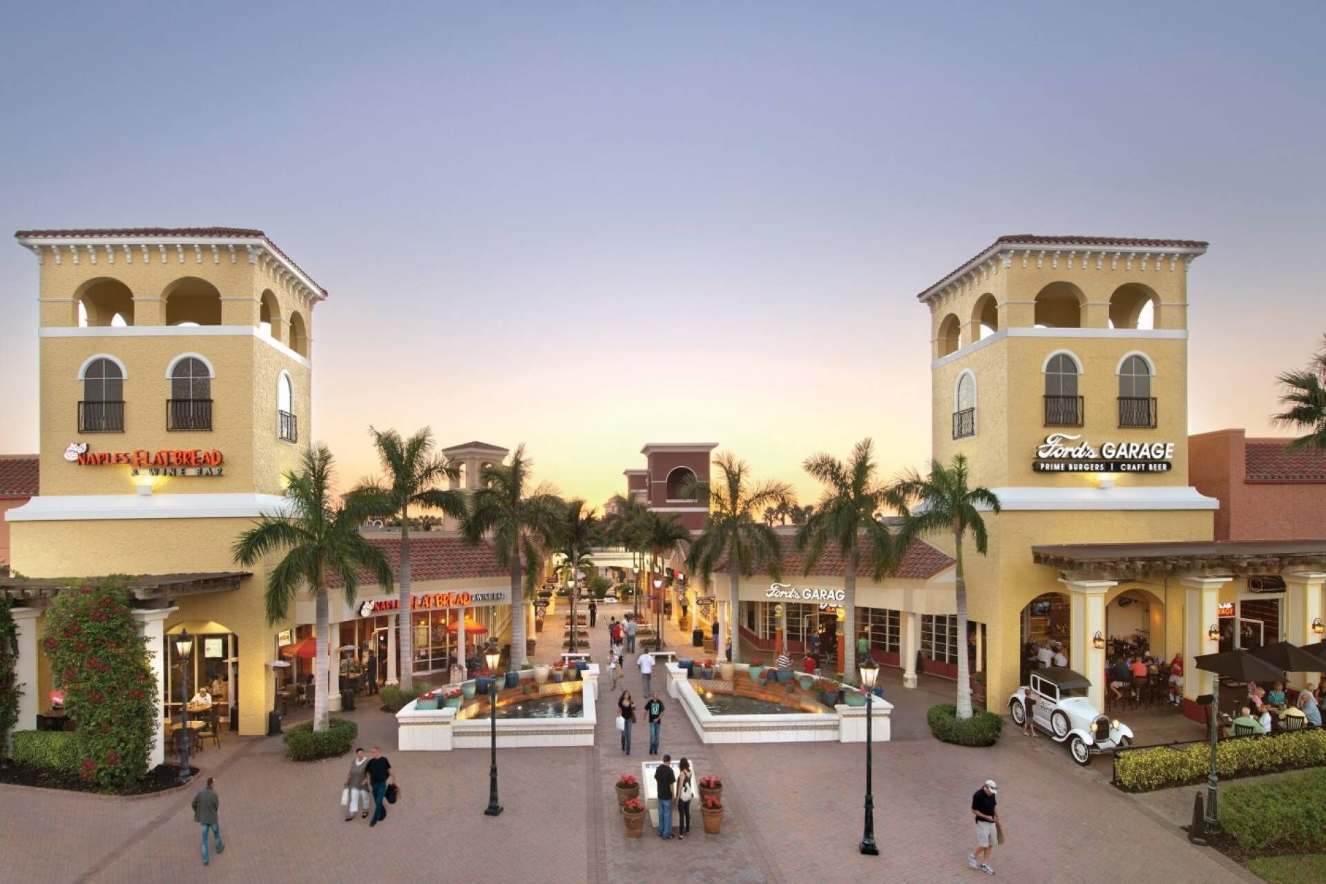 A shopping plaza with a central fountain, surrounded by palm trees and various storefronts including a flatbread and a garage.