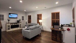 A modern living room with a grey sofa, wooden flooring, a TV mounted on the wall, and a small wooden side table with a lamp.