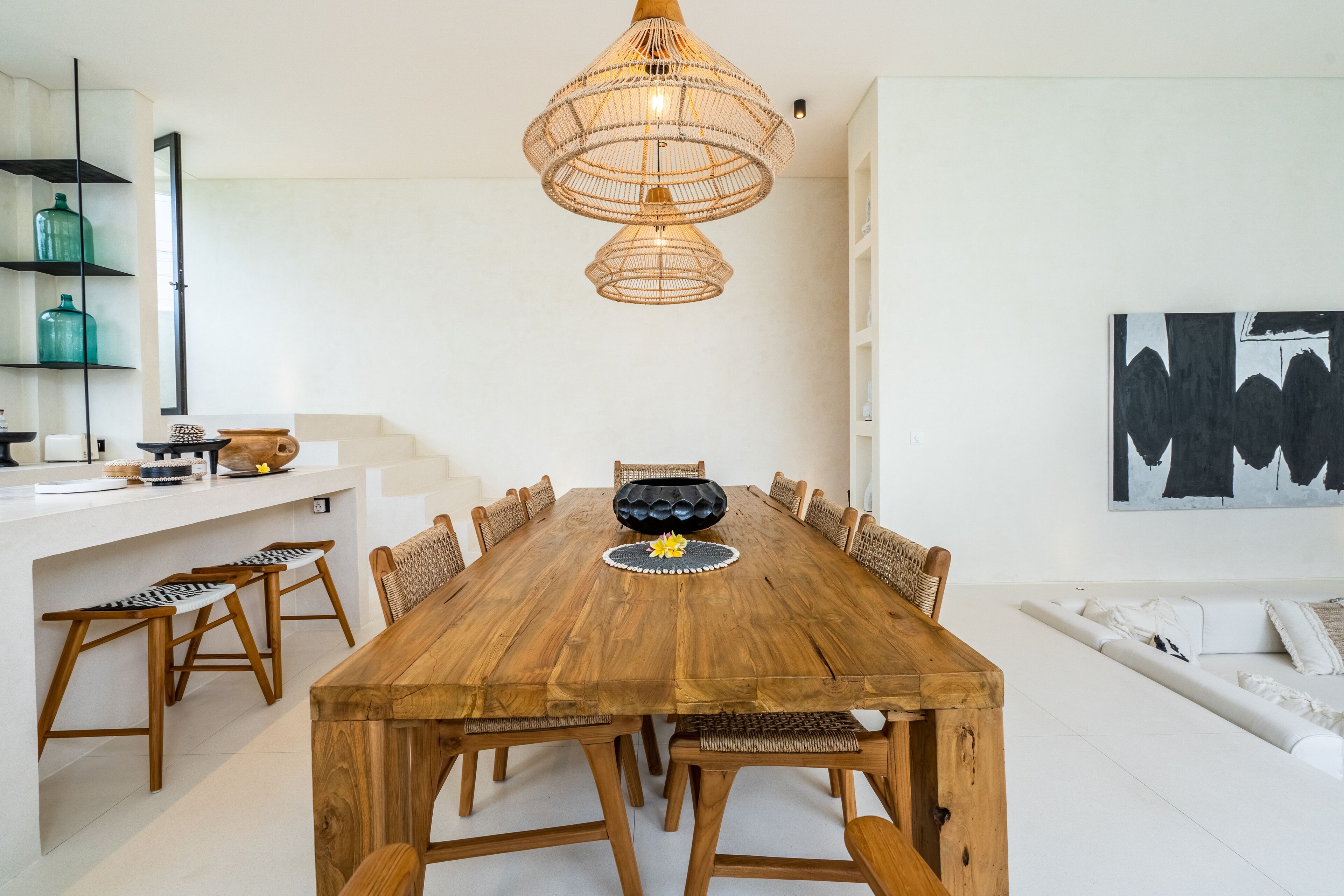 Een ruime eethoek met een houten tafel en stoelen, een hanglamp en een witte muur met een zwart-wit abstract schilderij.