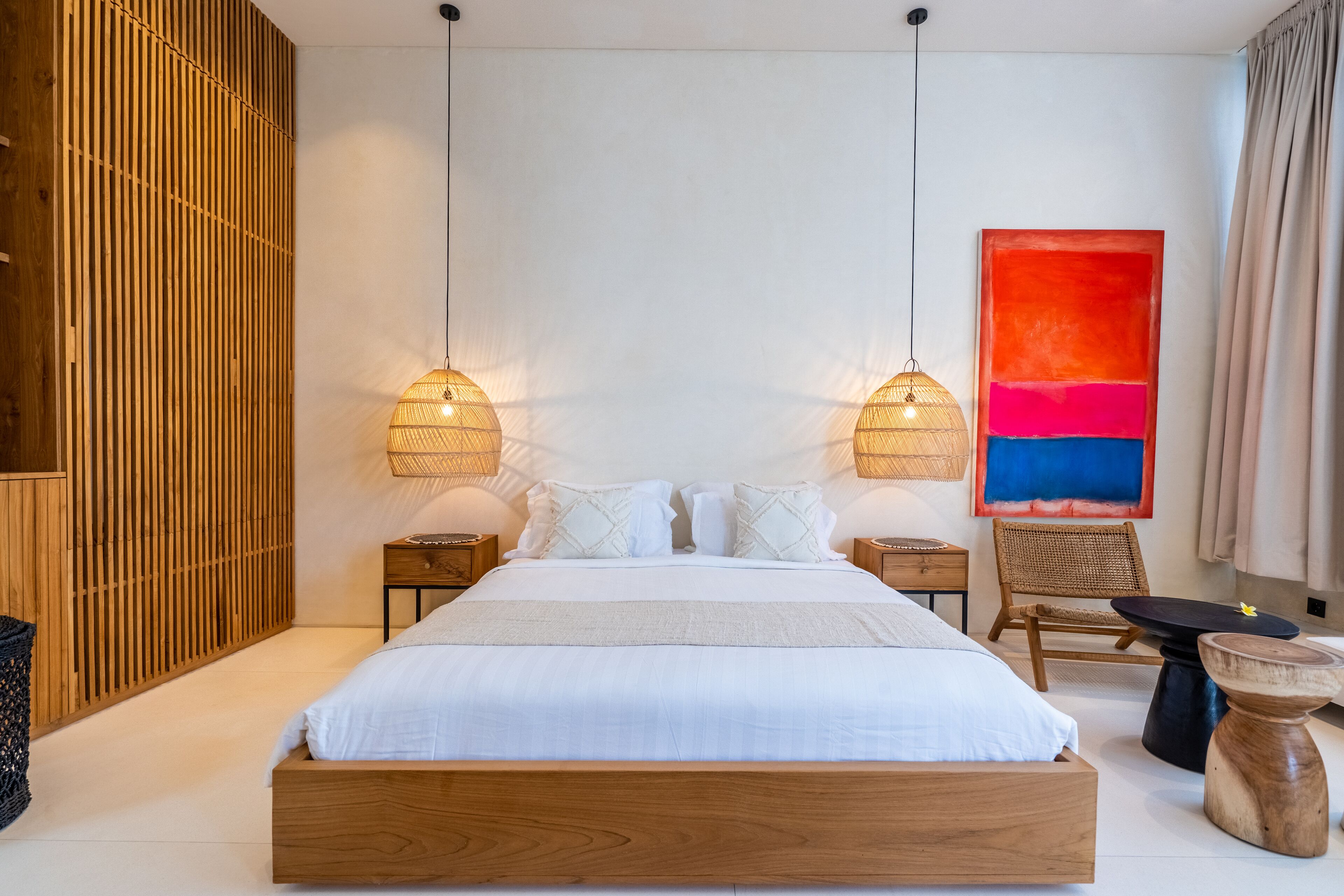 A modern bedroom with a wooden bed, two bedside lamps, a wicker chair, and a colorful abstract painting on the wall.