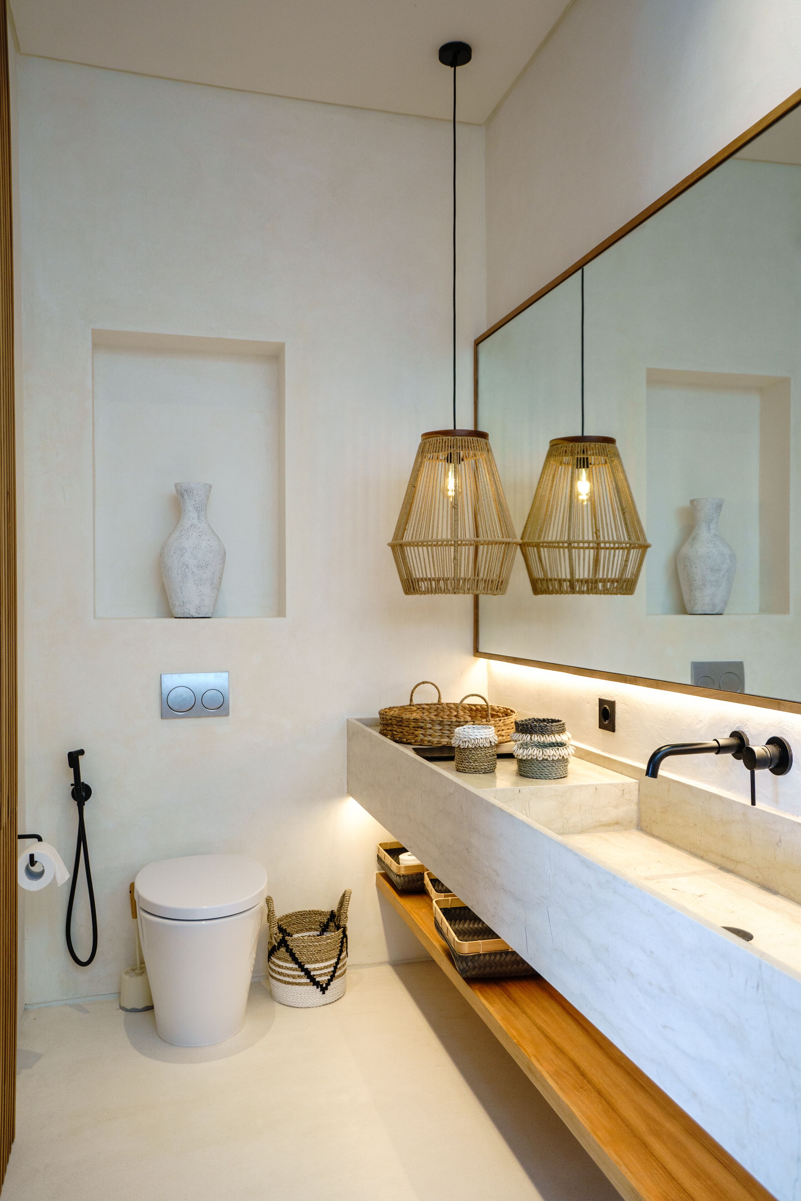 Une salle de bain moderne avec un grand miroir, deux luminaires suspendus, des toilettes et un meuble-lavabo doté de paniers et d’un évier.