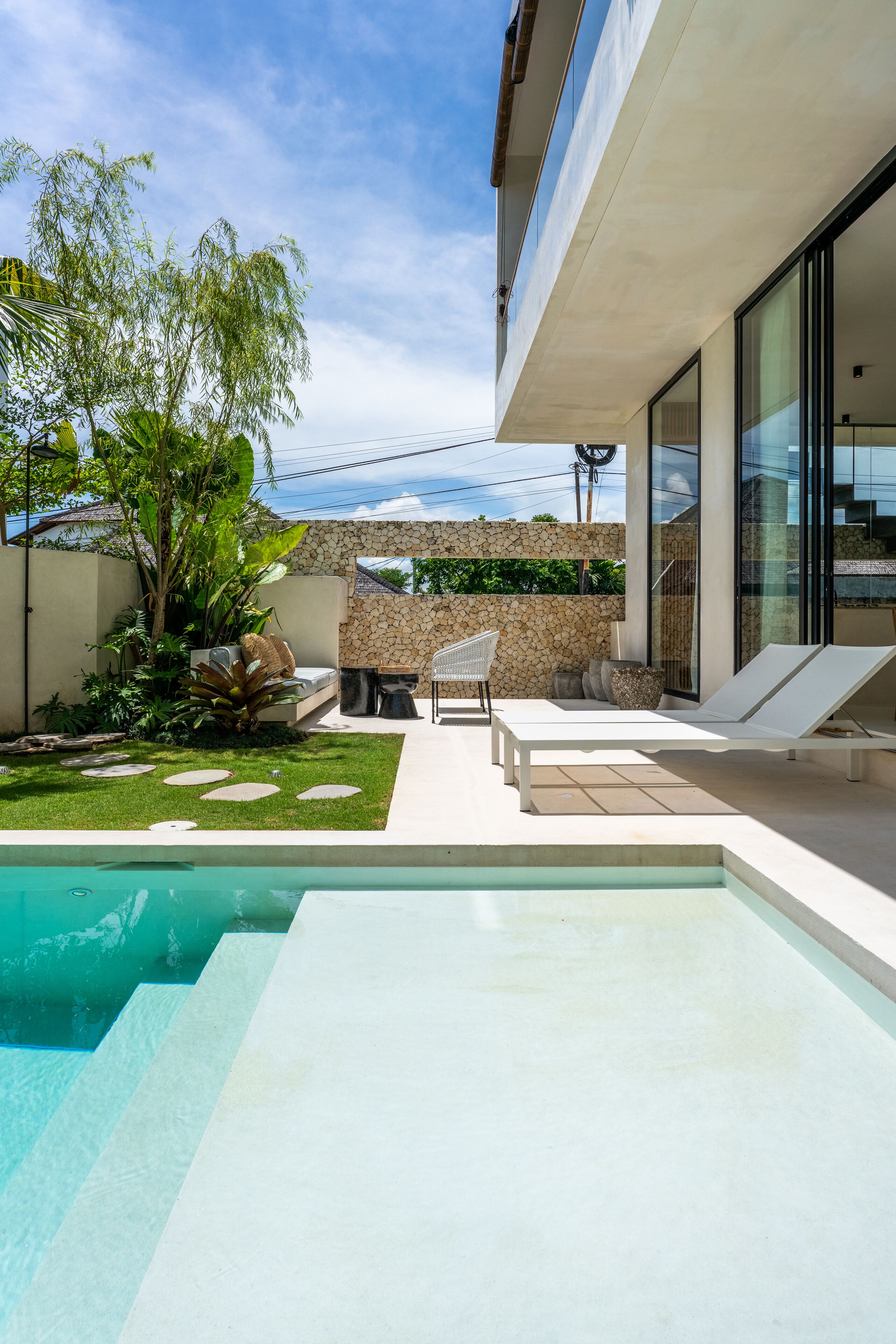 Un área de piscina moderna al aire libre con un muro de piedra, sillas de descanso y un pequeño jardín con plantas tropicales.