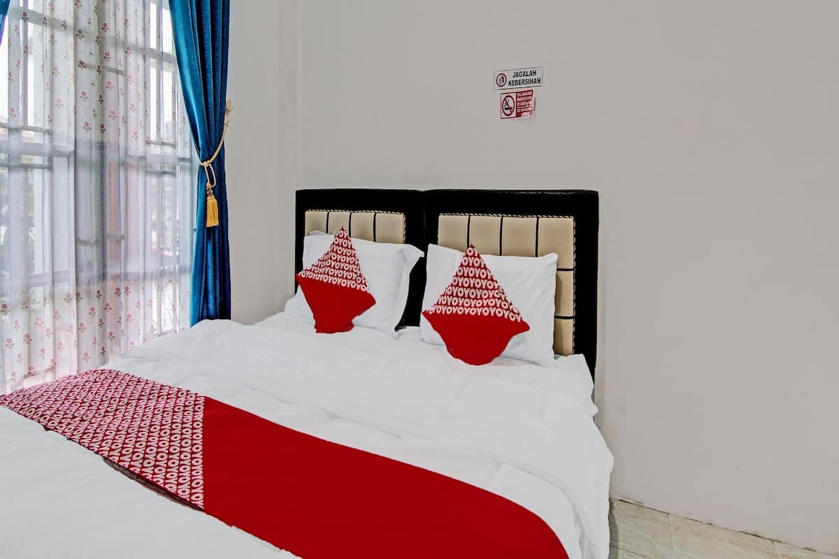A neatly made bed with white and red bedding, a headboard with a beige panel, and a window with floral curtains.