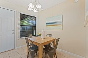 A dining area with a wooden table and four chairs, a window with blinds, a ceiling light fixture, and a painting on the wall.