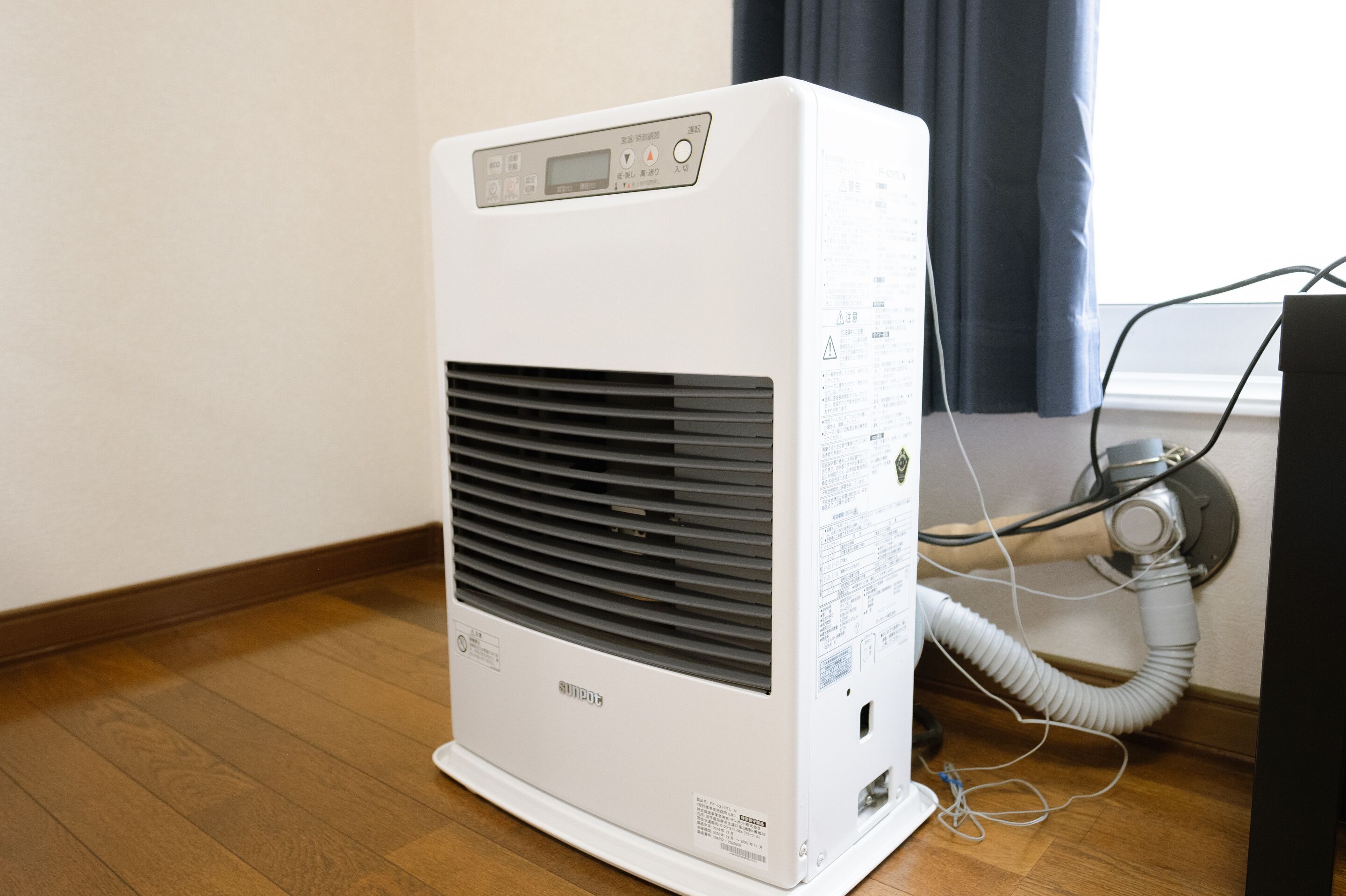 A white portable air conditioner with a digital display, placed on a wooden floor near a window with a curtain.