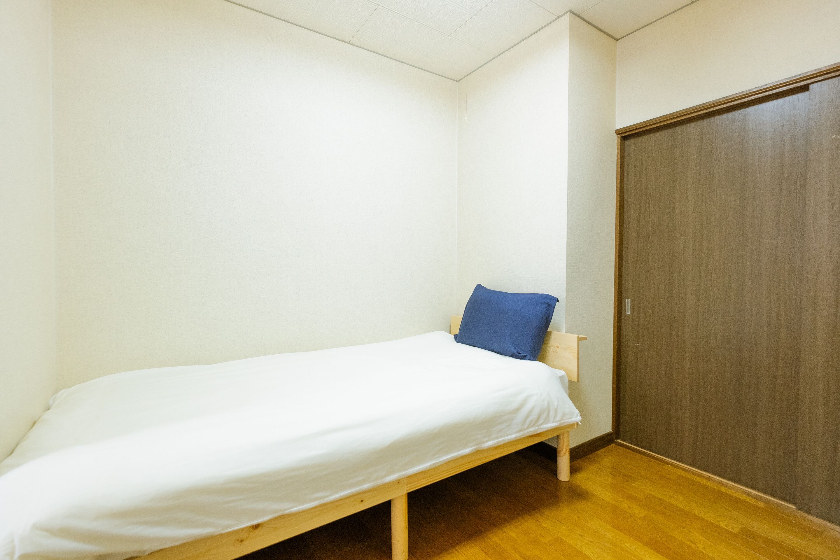 Ein Einzelbett mit weißer Matratze und blauem Kopfkissen in einem Zimmer mit Holzboden und einem Schrank aus Holz.