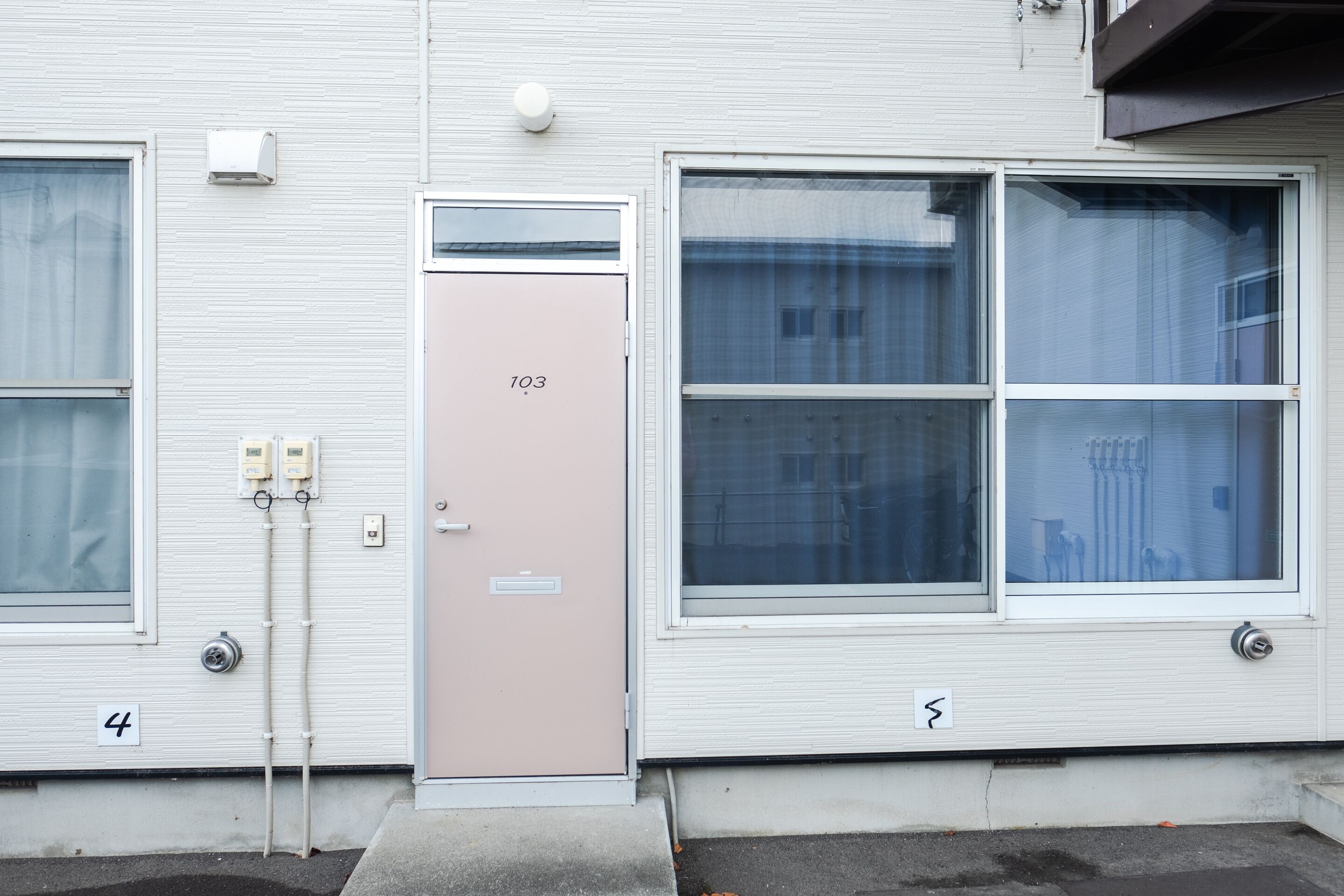 Un edificio con una puerta color beige con la etiqueta “103” y una ventana que ofrece vista a otro edificio al fondo.