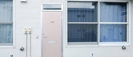 Un edificio con una puerta color beige con la etiqueta “103” y una ventana que ofrece vista a otro edificio al fondo.