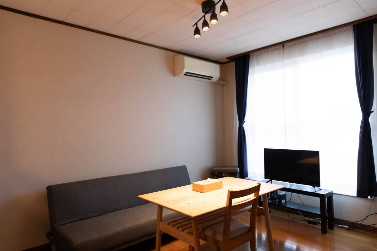 Pieni olohuone, jossa on puinen pöytä, harmaa sohva, litteänäyttöinen televisio ja ikkuna, jossa on verhot.