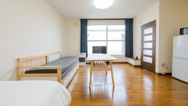 A room with a wooden bed, a small table, a desk, a window with curtains, and a refrigerator. - Furano Zone Ski Condo (Furano)