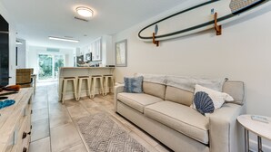 A modern living room with a beige sofa, a coffee table, and a surfboard mounted on the wall. - Fernandina Beach Paradise: Steps to Shore! (Fernandina Beach)