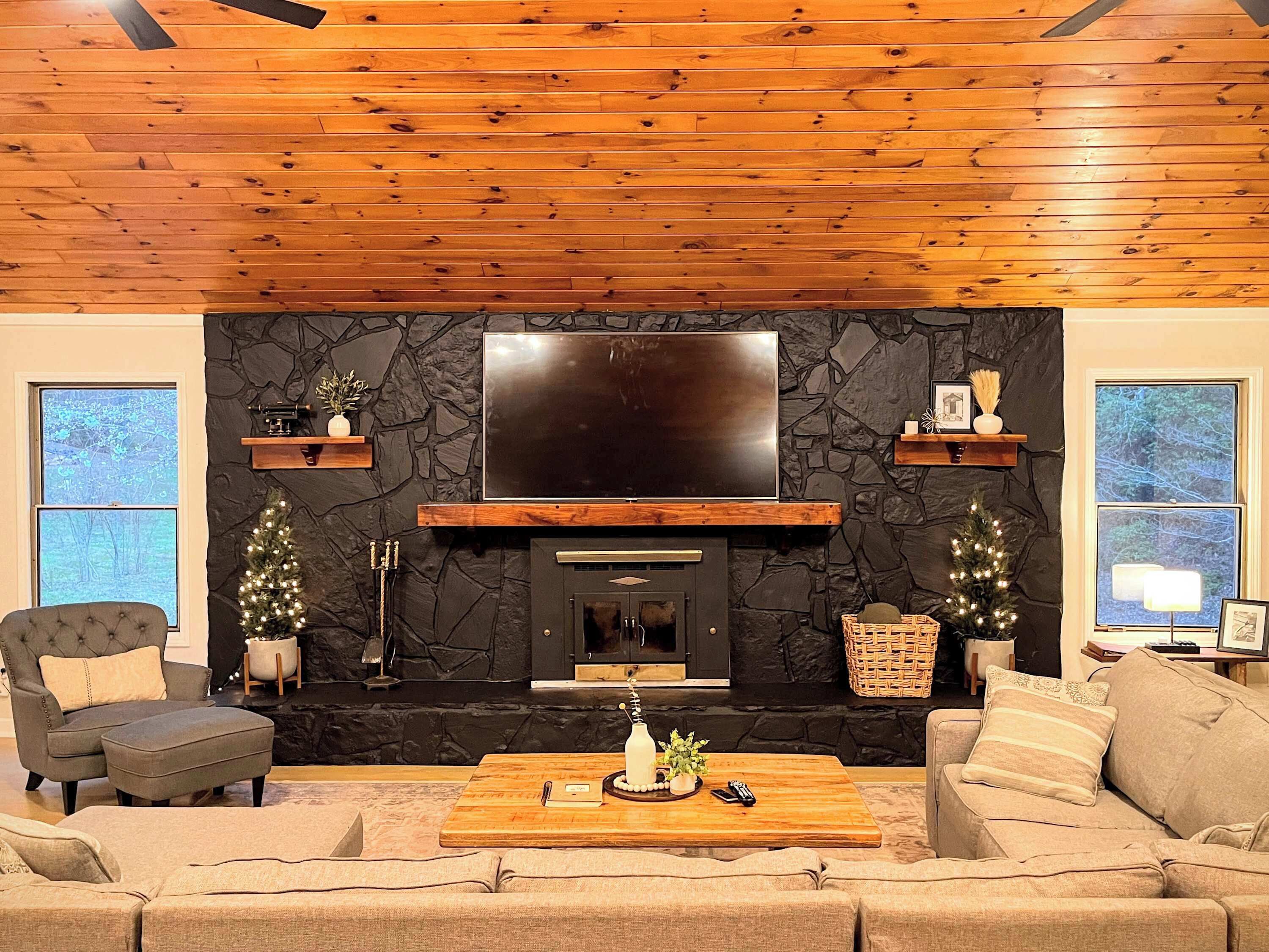 A living room with a stone fireplace, a flat-screen TV, and two windows.