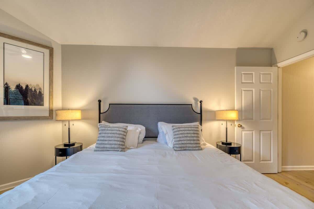 A bedroom with a large bed, two bedside tables with lamps, a framed picture on the wall, and a door.