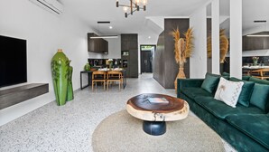Una sala moderna con un sofá verde, una mesa de centro de madera y un televisor a la izquierda. Se observa una cocina al fondo, junto con un comedor.