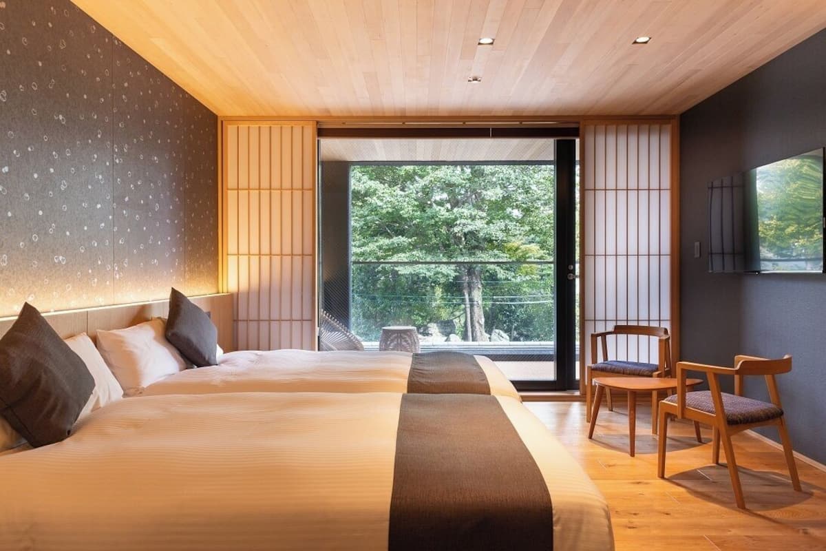 Une chambre d’hôtel moderne dotée d’un grand lit, d’un coin salon et offrant une vue sur les arbres grâce à une porte coulissante.