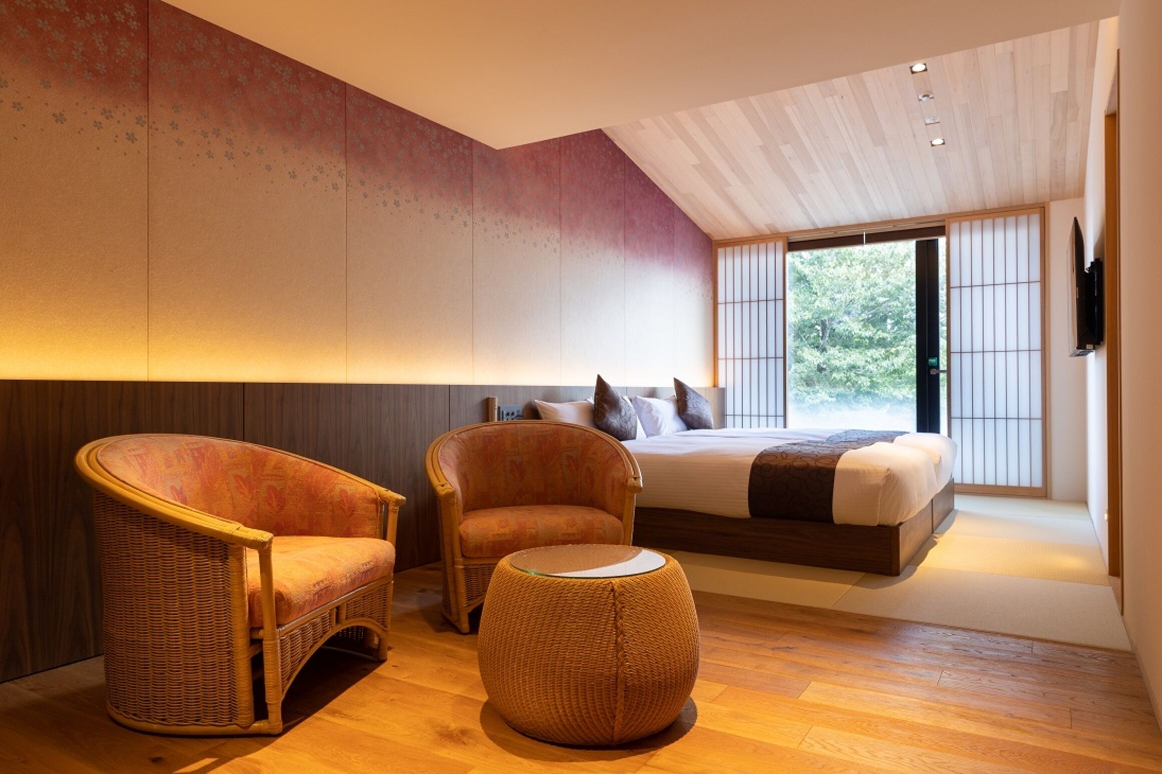 A modern bedroom with a large bed, two wicker chairs, a round ottoman, and a view of greenery through large windows.