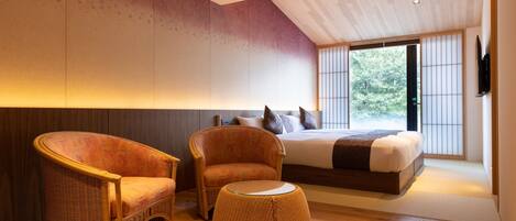A modern bedroom with a large bed, two wicker chairs, a round ottoman, and a view of greenery through large windows.