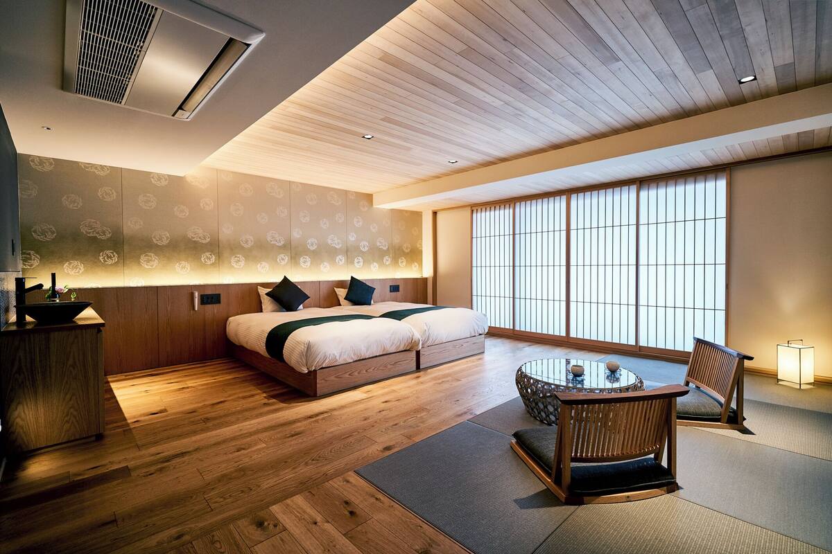 Une chambre d’hôtel moderne avec un plancher en bois, un grand lit avec des oreillers, une table de chevet et une vue sur l’extérieur grâce à de grandes fenêtres.