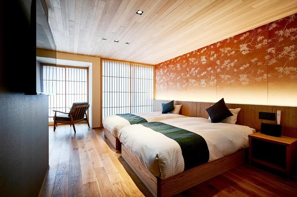 A hotel room with a wooden ceiling, two beds with green and white bedding, a wooden nightstand, a chair, and a large window with vertical blinds.