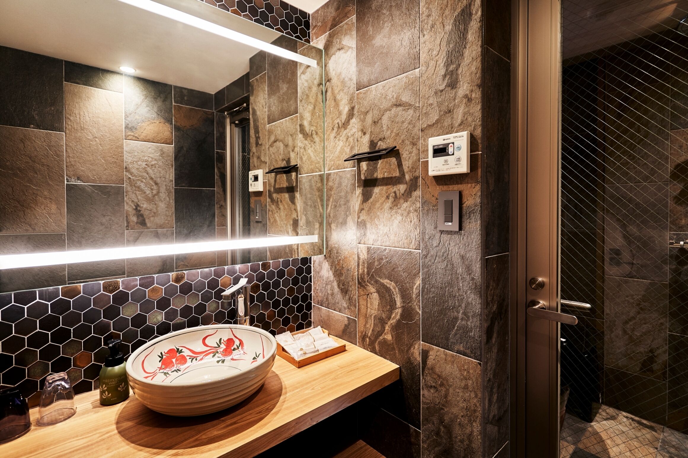 Een badkamer met een houten wastafelmeubel, een rond wastafelblad met decoratieve kom, een spiegel met ingebouwde verlichting en een douchecabine met glazen deur.