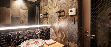 Een badkamer met een houten wastafelmeubel, een rond wastafelblad met decoratieve kom, een spiegel met ingebouwde verlichting en een douchecabine met glazen deur.