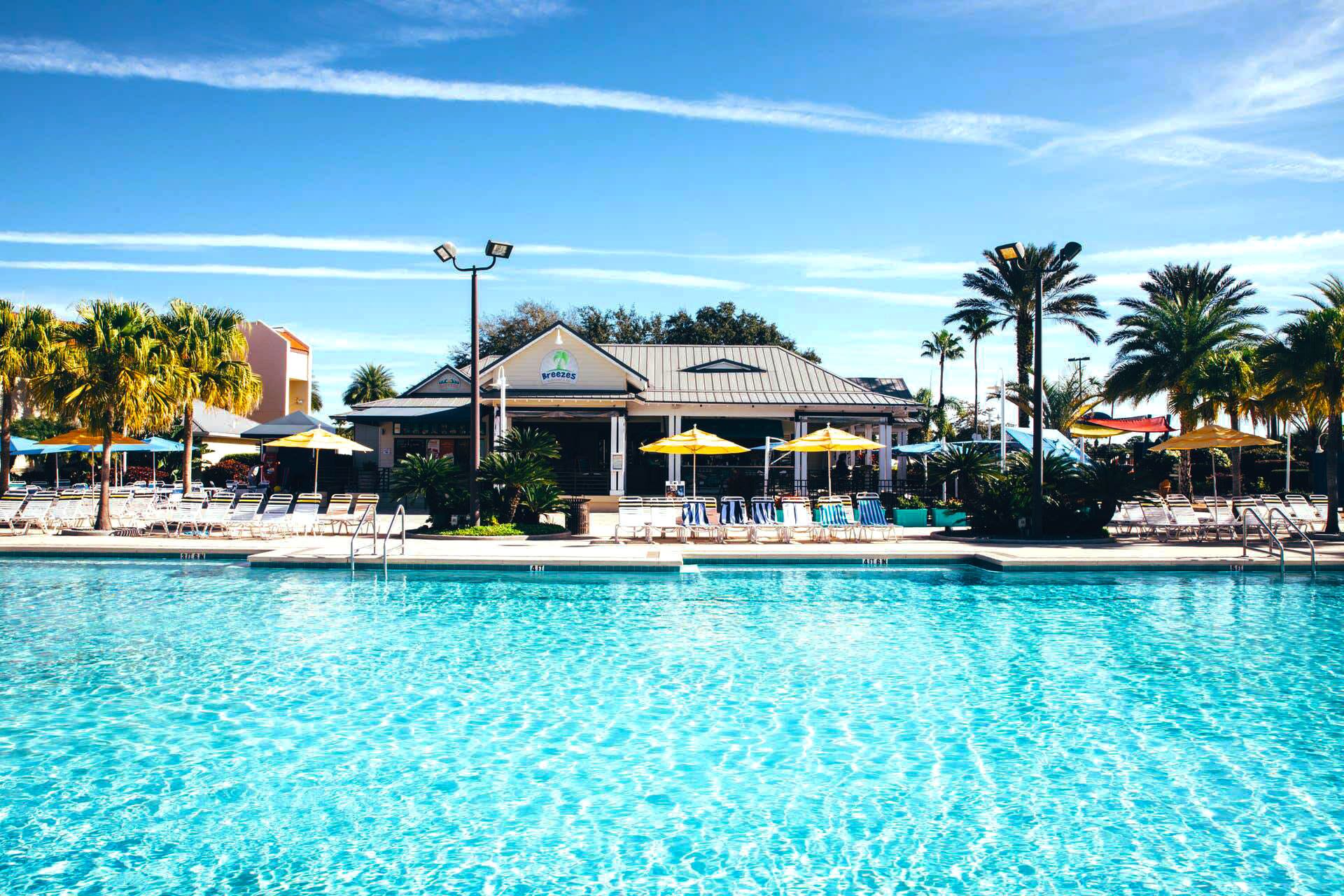 Une piscine avec des chaises longues et des parasols, un bâtiment sur lequel on peut lire l’inscription « Breezes », et des palmiers.