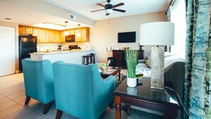 A modern living room with teal chairs, a coffee table, and a TV. There is a kitchen in the background with wooden cabinets and a microwave.