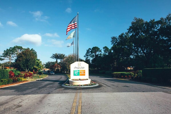 Señal de Orange Lake Resort & Spa con un asta de bandera, un parterre circular y una carretera que lleva a la propiedad.