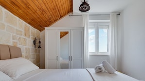 Een slaapkamer met een stenen muur, houten plafond, een bed met witte lakens, een kledingkast en een raam met gordijnen.