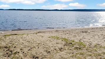 A sandy beach with a calm lake and a forested shoreline in the distance.