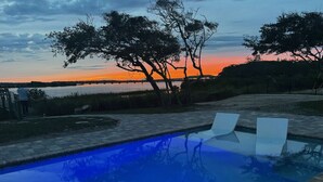 Une piscine avec deux chaises longues, entourée d’arbres et offrant une vue sur le coucher de soleil se reflétant sur une étendue d’eau.