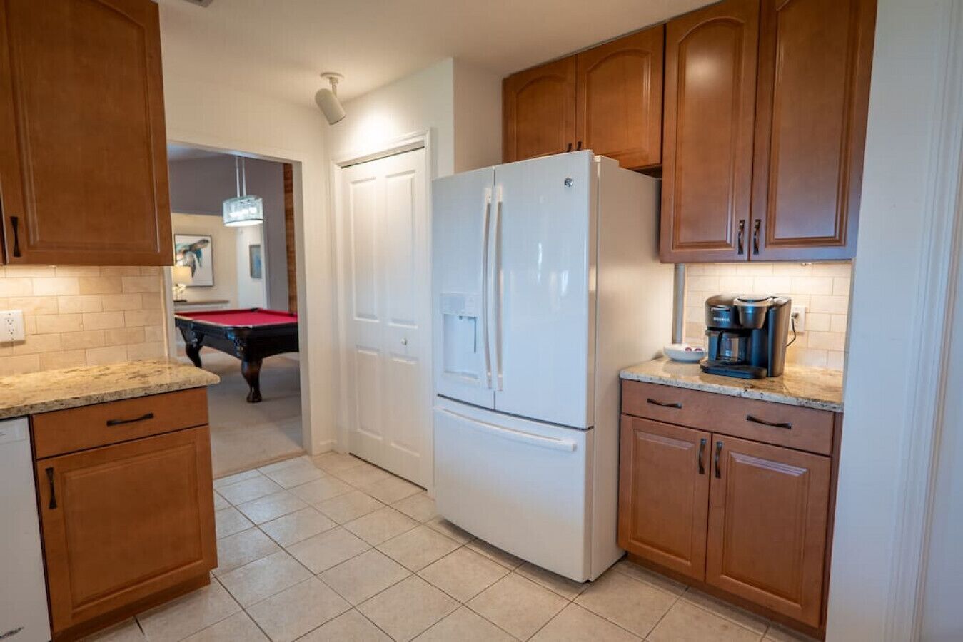 A kitchen with wooden cabinets, a white refrigerator, a coffee maker, and a pool table in the background.
