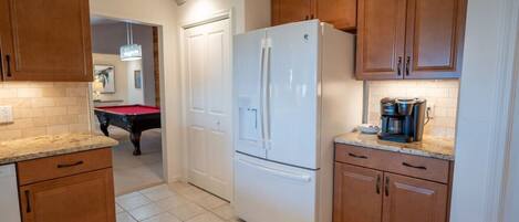 A kitchen with wooden cabinets, a white refrigerator, a coffee maker, and a pool table in the background.