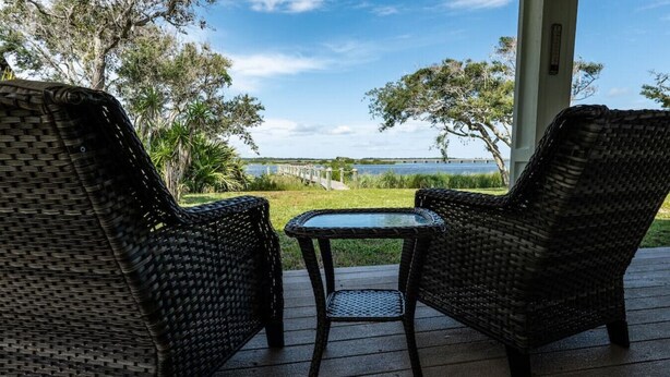 Deux fauteuils en osier et une petite table sur une terrasse en bois, avec vue sur un pont et sur l’eau.