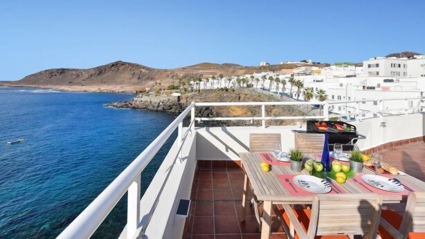 Um terraço com mesa posta para refeições, com vista para uma cidade costeira com edifícios brancos e céu azul claro.