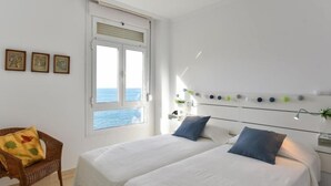 Quarto com duas camas, janela com vista para o mar, uma cadeira e luminárias na parede.