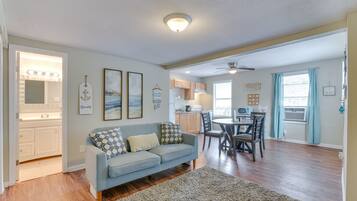 A living room with a sofa, a rug, a dining area with chairs and a table, a kitchen with cabinets and appliances, and a ceiling fan.