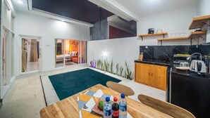 A modern kitchen with a black countertop, wooden cabinets, and a built-in stove. There is a dining area with a wooden table and chairs, and a small pool in the center. - MODERN COZY PRIVATE 2BR VILLA WITH ROOF TOP SUNSET VIEW (Bali)