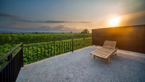 A balcony with a wooden lounge chair, a black metal railing, and a view of greenery and the sunset. - MODERN COZY PRIVATE 2BR VILLA WITH ROOF TOP SUNSET VIEW (Bali)