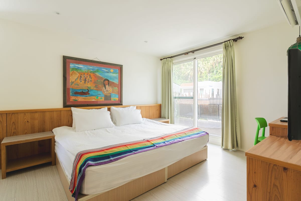 A bedroom with a wooden bed, a colorful blanket, a painting on the wall, and a view through the window.