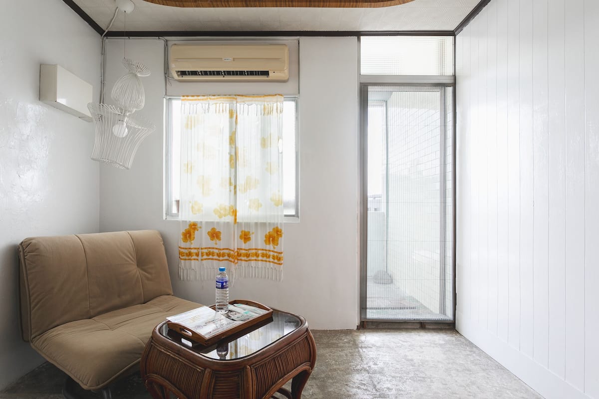 A room with a beige sofa, a wooden table with a tray and a bottle, a decorative hanging light, an air conditioner, and a window with a curtain.