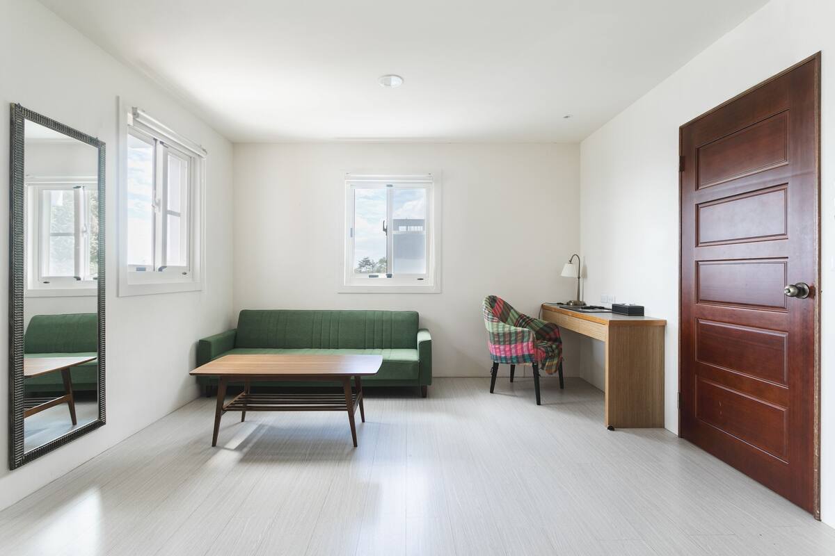 A room with a green sofa, a wooden table, a chair, a desk, a mirror, and a door.