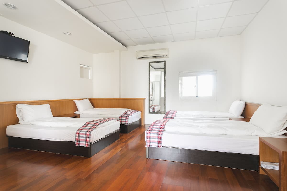 A hotel room with three beds, wooden flooring, a TV, and an air conditioning unit.